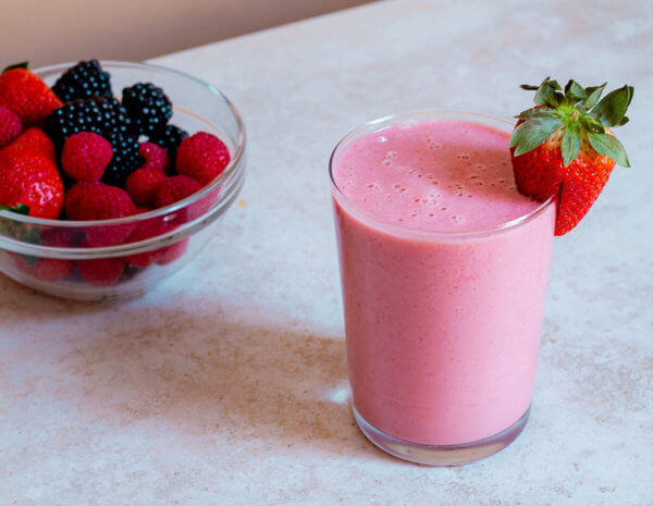 A glass of oat and almond butter smoothie with strawberry along side a bowl of mixed berries