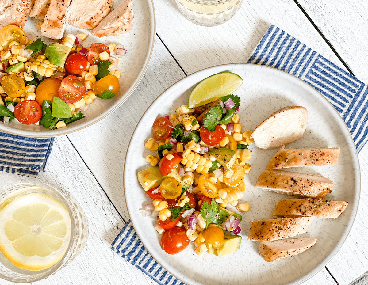 A top down view of a plate of grilled chicken with corn, avocado & tomato salad